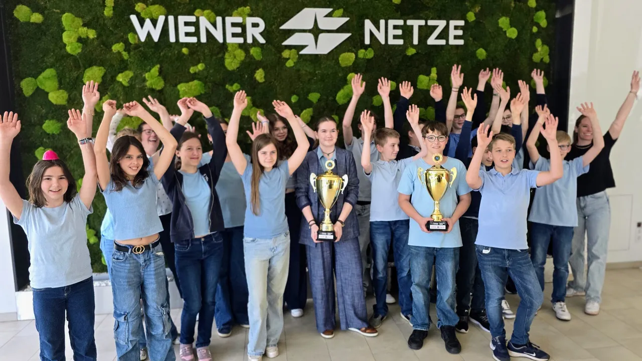 Eine ausgezeichnete Schulklasse jubelt mit erhobenen Armen und zwei Pokalen vor der Mooswand der Wiener Netze