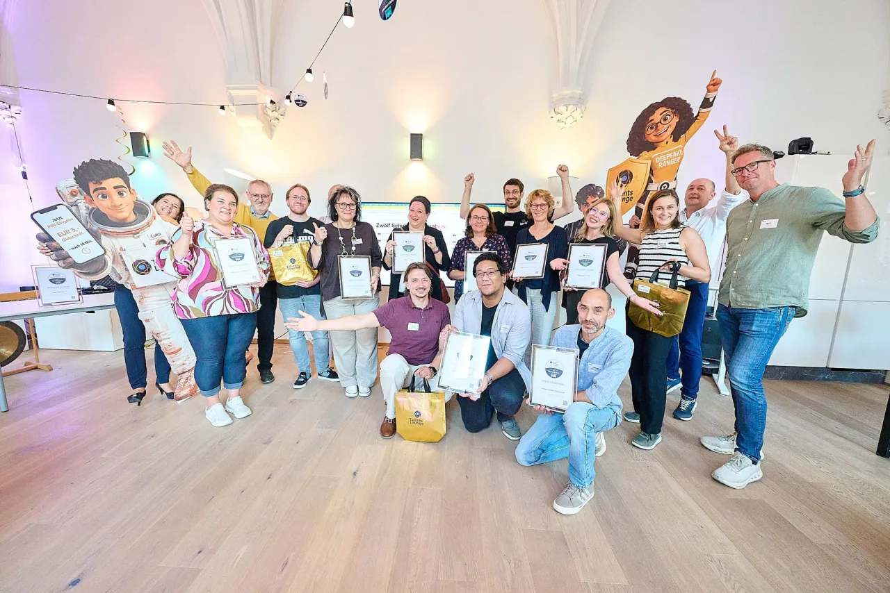 Gruppenfoto der ausgezeichneten Wiener Lehrkräfte mit ihren gerahmten Urkunden im Saal des DaVinciLab, viele jubeln mit erhobenen Armen, daneben die lebensgroßen Figuren