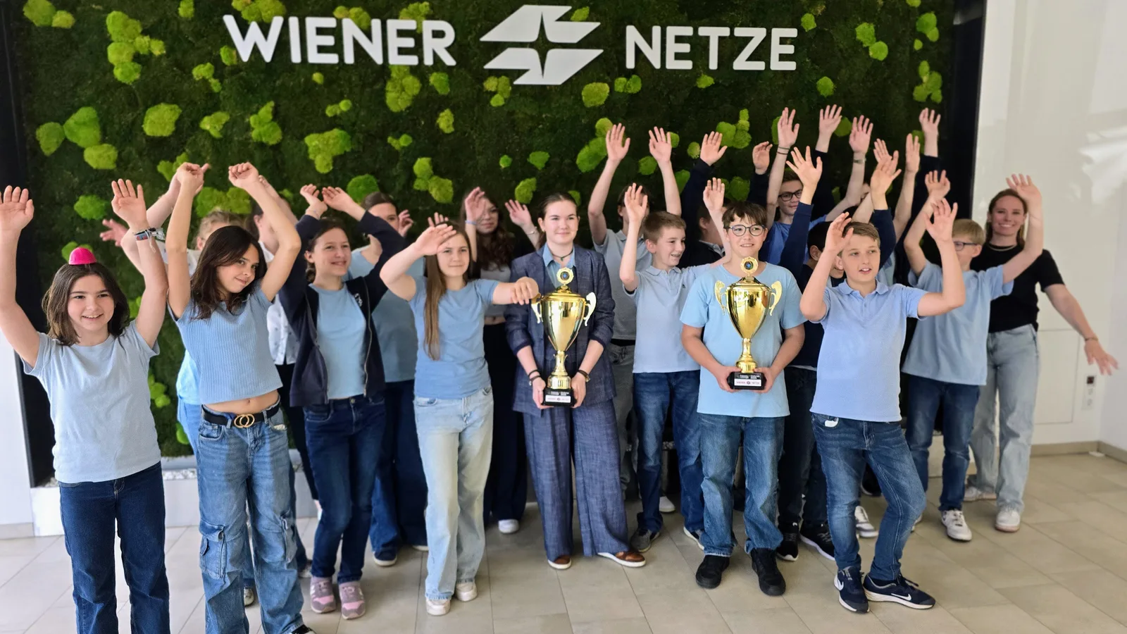 Jubelnde Schulklasse mit Pokalen vor der begrünten Medienwand der Wiener Netze