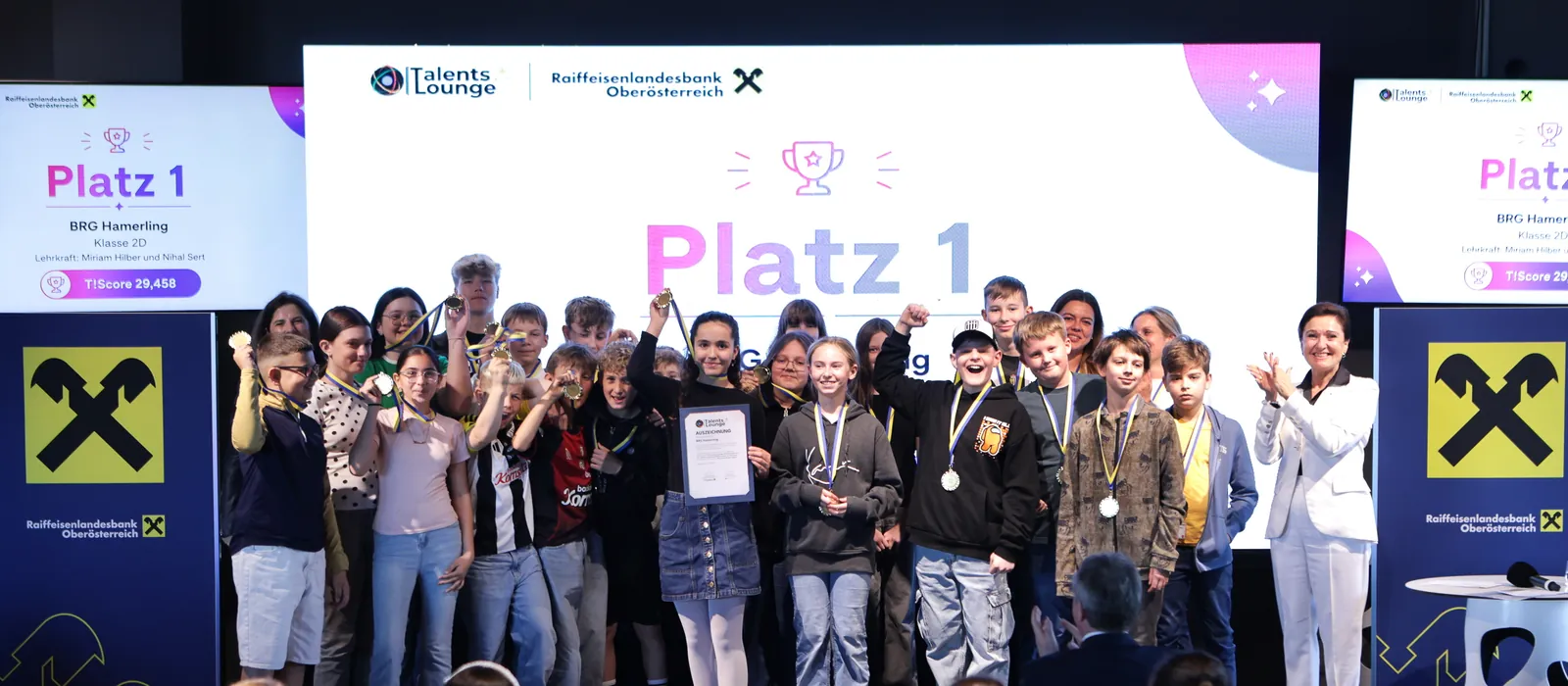 Die 2D des BRG Hamerling feiert Platz 1 der RLB OÖ TalentsLounge Challenge auf der Bühne der Raiffeisen Arena, rechts applaudiert Sigrid Burkowski